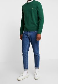 Mann in grünem Rundhalsausschnittpullover, blauen hochgekrempelten Jeans, weißen Socken und weißen Sneakers steht vor einem weißen Hintergrund.