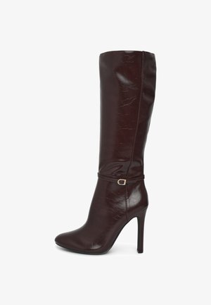 Botte en cuir marron jusqu'aux genoux avec une finition brillante, bout pointu et talon aiguille de 10 cm. Présente une sangle au niveau de la cheville avec un accent de boucle en or.