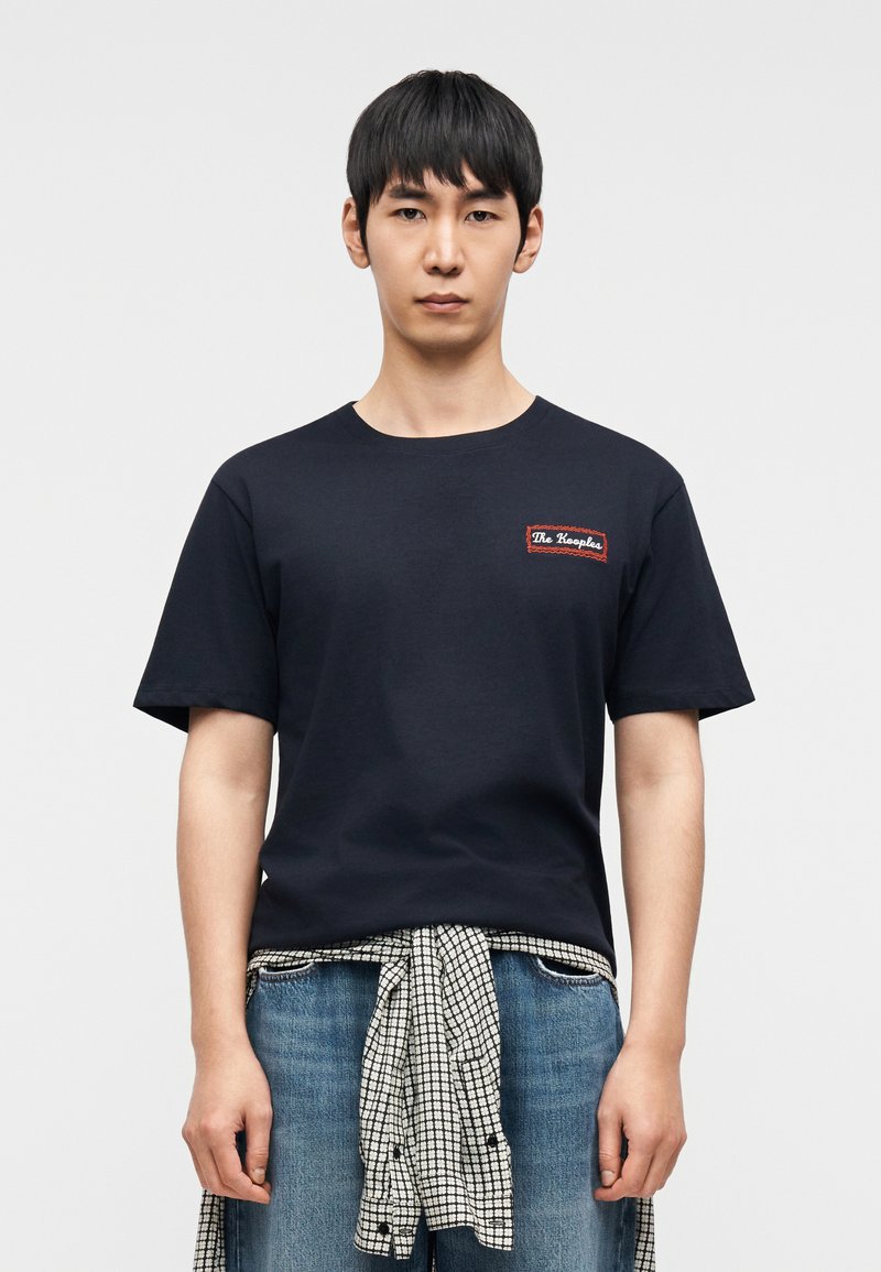 Navy blue cotton t-shirt with a round neckline and short sleeves. Features a red embroidered patch on the chest. Worn with checkered waist tie.