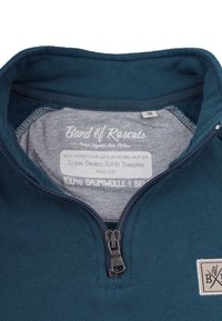 Økologisk bomulds zip-up sweatshirt i mørk tyrkis. Har en grå inderside, en lynlås lukning og et logo patch på brystet.