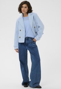Femme portant une veste double boutonnage texturée bleu clair, un pull assorti, un jean bleu à jambes larges et des chaussures noires, posant avec une main sur la hanche.