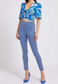 Vrouw in blauwe high-waisted broek met zilveren knopen, een blauw-wit cropped shirt met palmbladpatroon, en blauwe hoge hakken.