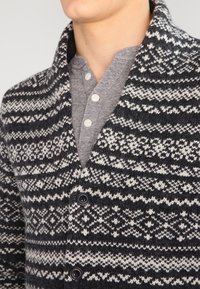 Cárdigan de punto con un patrón geométrico en blanco y negro, que presenta un cuello chal y tres botones oscuros sobre una camiseta henley gris.