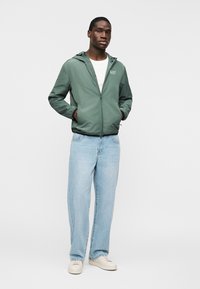 Mand stående med hænderne i lommerne iført grøn hættetrøje, hvid skjorte, lyseblå jeans og hvide sneakers mod ensfarvet baggrund.