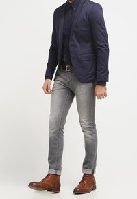 Marineblazer over een donker shirt, gecombineerd met lichtgrijze jeans. Bruine leren enkellaarzen, iets opgerolde mouwen en knoopaccenten op het shirt.