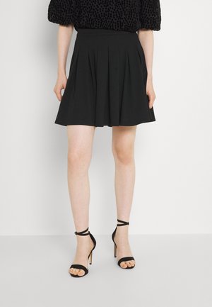 Kvinde iført sort plisseret nederdel, sorte hælede sandaler med ankelremme og sort mønstret kortærmet top, stående mod hvid baggrund.