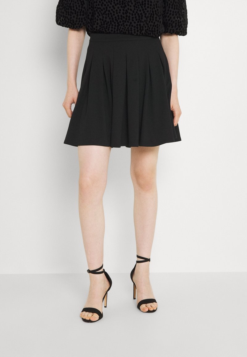 Femme portant une jupe plissée noire, des sandales à talons noires avec brides autour de la cheville et un haut à manches courtes noir à motifs, se tenant devant un fond blanc.