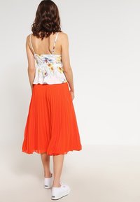 Květinový peplum top s tenkými ramínky spárovaný s plisovanou sukní střední délky v oranžové barvě. Bílá teniska dotváří outfit.