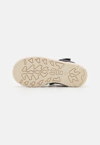 Musta sandaali, jossa on teksturoitu musta päällinen, säädettävä nauha ja beige kumipohja, jossa on yksityiskohtainen pito.