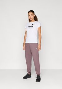 Biela tričko s krátkym rukávom a čiernym logom Puma, v kombinácii so svetlo fialovými teplákmi s malým logom. Oblečené s čiernymi športovými topánkami.