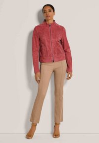 Rosa Wildlederjacke mit hohem Kragen und Reißverschluss vorne; verfügt über seitliche Reißverschlusstaschen und strukturierte Einsätze. Das Modell trägt beigefarbene, eng anliegende Hosen und hellbraune Absätze.