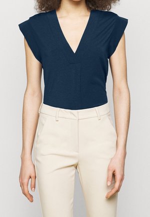 Vrouw draagt een marineblauw mouwloos V-hals topje dat is ingestopt in een beige, hooggetailleerde broek met riemlussen.