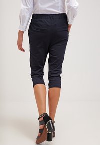 Pantalons cropped navy avec une taille élastique et deux poches arrière, associés à des sandales à talons hauts noires avec des accents dorés.