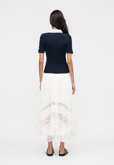 Γυναίκα που στέκεται με την πλάτη γυρισμένη, φορώντας κοντομάνικη μπλούζα σε μπλε σκούρο ριγέ με λευκό γιακά και λευκή πλισέ φούστα με δαντελένιες λεπτομέρειες και βολάν στο τελείωμα.