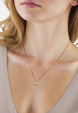 Nærbillede af en kvindes nakke og bryst iført en fin halskæde med en gylden stjerneformet vedhæng, med lysebrunt hår og en beige top.