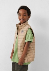 Jonge jongen lacht, draagt een beige doorgestikt vest over een groen shirt met korte mouwen, staand tegen een effen witte muur.