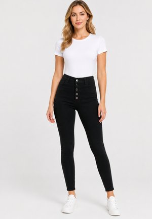 Kvinde med langt krøllet hår bærer en tætsiddende hvid kortærmet top, højtaljede sorte skinny jeans med knapper foran og hvide sneakers.