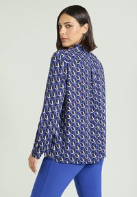 Bluzka z długim rękawem z geometrycznym wzorem w odcieniach niebieskiego i białego na ciemnym tle, o luźnym kroju z plisą na plecach.