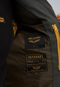 Siva jakna z notranjostjo in rumenim zadrgo. Ima tekstilne oznake: "PME LEGEND" in "YETCRAFT" na črni podlagi z zlatim napisom.