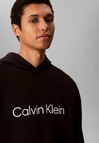 Mladi moški s kratkimi kodrastimi lasmi, ki nosi črn pulover s kapuco z napisom „Calvin Klein“ na sprednji strani, na enobarvnem sivem ozadju.