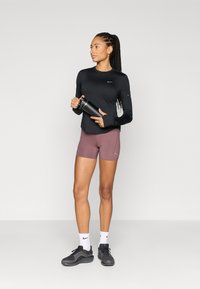 Haut noir à manches longues, short violet et baskets noires. Tenant une bouteille d'eau noire. Design minimaliste avec logos Nike sur les vêtements.