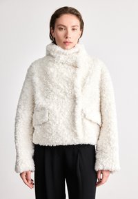 Biała kurtka z sztucznego futra z wysokim kołnierzem, skróconym krojem, bocznymi kieszeniami i długimi rękawami, zestawiona z czarnymi spodniami.