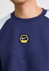 Námornícky modrý mikina so zaoblenou čiernou a žltou nášivkou Nike. Obsahuje svetlomodré ramenné panely a rebrovaný výstrih. Hladká textúra.