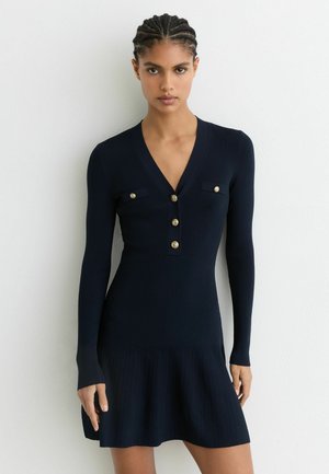 Marineblauwe geribde gebreide jurk met een aangesloten lijfje, lange mouwen, V-hals, gouden knoopaccenten, twee borstzakken en een uitlopende rok.