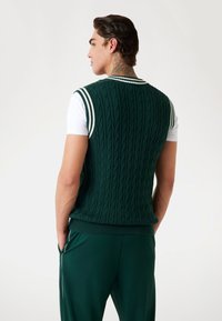 Zelený pletený vest s bílým pruhovaným ribbed límcem, spárovaný s bílým tričkem. Materiál vypadá měkce a texturovaně.