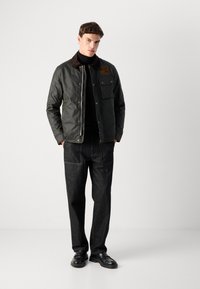 Veste en coton ciré vert foncé avec un col en velours côtelé marron, boutons sur le devant, poche poitrine, associée à un jean en denim foncé et des chaussures noires.
