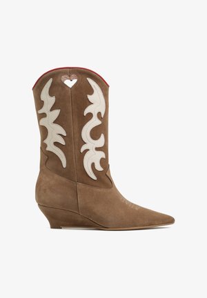 Brązowy zamszowy kowbojski but z białymi wzorami przypominającymi płomienie, wycięciem w kształcie serca blisko góry, spiczastym noskiem i niskim, nachylonym obcasem klinowym.