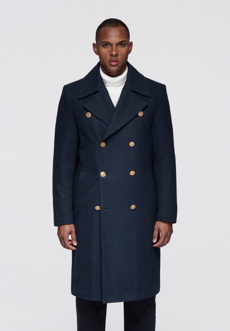 Marineblå uld dobbeltradet jakke med store gyldne knapper, brede revers, sidelommer og en skræddersyet silhuet. Båret over en hvid rullekrave.