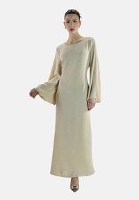 Lange beige jurk met een vierkante halslijn, brede klokmouwen en een gestructureerde stof. Gecombineerd met puntige schoenen voorzien van een decoratief accent.