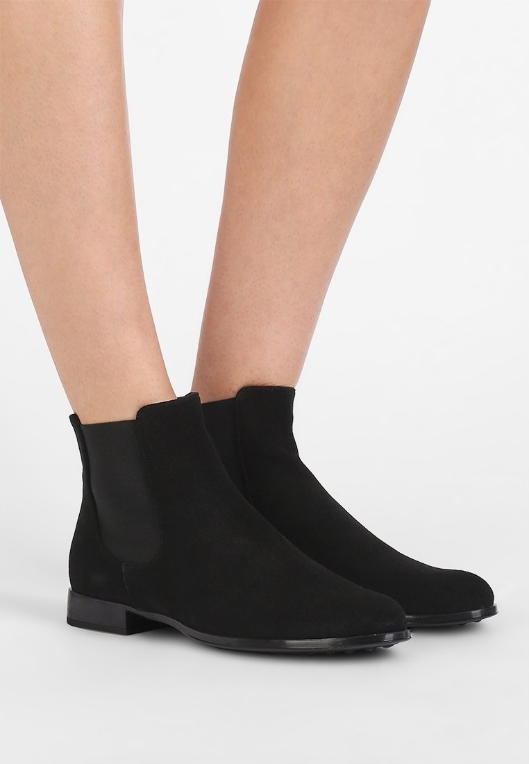 Bottes Chelsea en daim noir avec panneaux latéraux élastiques portées sur des jambes nues, présentées sur un fond blanc uni.