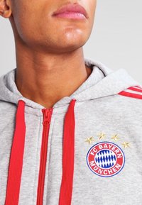 Hoodie gris zippé avec des accents rouges, logo brodé du FC Bayern München et trois étoiles dorées au-dessus du logo sur la poitrine.
