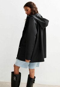 Wodoodporna czarna kurtka z kapturem, luźny krój, zatrzaski i kieszenie z przodu, połączona z jasnoniebieskimi szortami dżinsowymi i czarnymi butami.