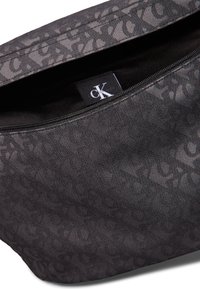 Bolso de mano de tela negra de Calvin Klein con un sutil patrón de logotipos en relieve, cierre de cremallera y una superficie lisa y texturizada.