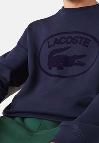 Laivastonsininen crewneck-collegepaita pehmeästä kankaasta, jossa on kohokuvioitu Lacoste-logo ja keskellä alligaattorikuvio. Resorit hihansuissa ja helmassa.