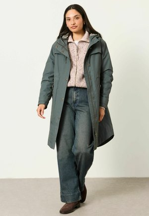 Frau mit langen dunklen Haaren trägt einen langen grünen Mantel, pinken gestrickten Reißverschluss-Pullover, weite blaue Jeans und braune Stiefeletten und geht vorwärts.