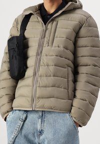 Nafouklá olivově zelená bunda s horizontálním prošíváním, kapucí a předním zipem. Přes rameno je přehozena malá černá taška.