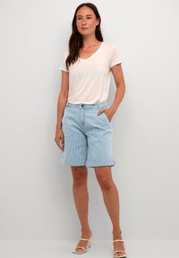T-shirt rosa chiaro a V abbinata a pantaloni corti blu a righe con risvolto. Il modello è in sandali con lacci, con le mani nelle tasche. Design semplice.