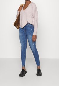 Kvinna iklädd en ljusrosa V-ringad tröja, blå slitna skinny jeans, svarta slip-on skor och bär en brun fuskpäls axelväska.