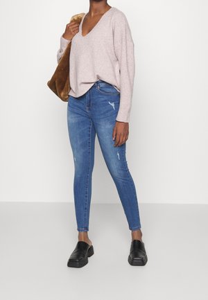 Kvinna iklädd en ljusrosa V-ringad tröja, blå slitna skinny jeans, svarta slip-on skor och bär en brun fuskpäls axelväska.