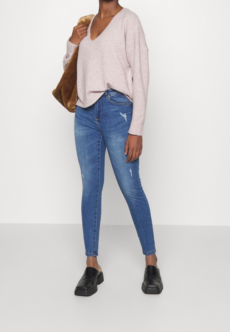 Kvinna iklädd en ljusrosa V-ringad tröja, blå slitna skinny jeans, svarta slip-on skor och bär en brun fuskpäls axelväska.