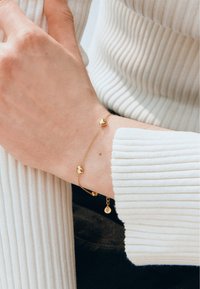 Bracciale in oro con tre piccoli ciondoli a forma di cuore, caratterizzato da una delicata catena. In primo piano, una mano su un tessuto leggero e testurizzato.