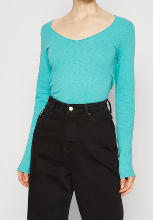 Personne portant un haut ajusté turquoise à manches longues avec un large col en V, rentré dans un pantalon noir ample taille haute.