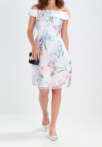 Sukienka z odkrytą ramieniem, w kwiaty, z poziomymi paskami w jasnych odcieniach. Posiada intensywne wzory kwiatowe w kolorach różowym, niebieskim i zielonym. W zestawieniu z srebrnymi szpilkami.