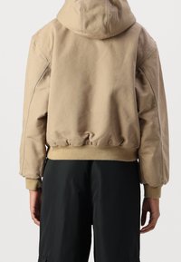 Beige hoodie met een relaxed fit, geribde manchetten en zoom. Gladde stof met een minimalistisch ontwerp, weergegeven van achteren tegen een lichte achtergrond.