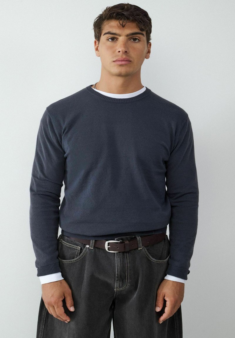 Donkerblauwe crewneck sweater met geribde manchetten, gedragen over een wit shirt, in combinatie met zwarte jeans en een bruine riem. Zachte textuur, casual pasvorm.