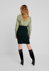 Olivgrön blus med långa ärmar, ihop med en mörkgrön texturerad pennkjol. Modellen bär knähöga svarta klackstövlar.